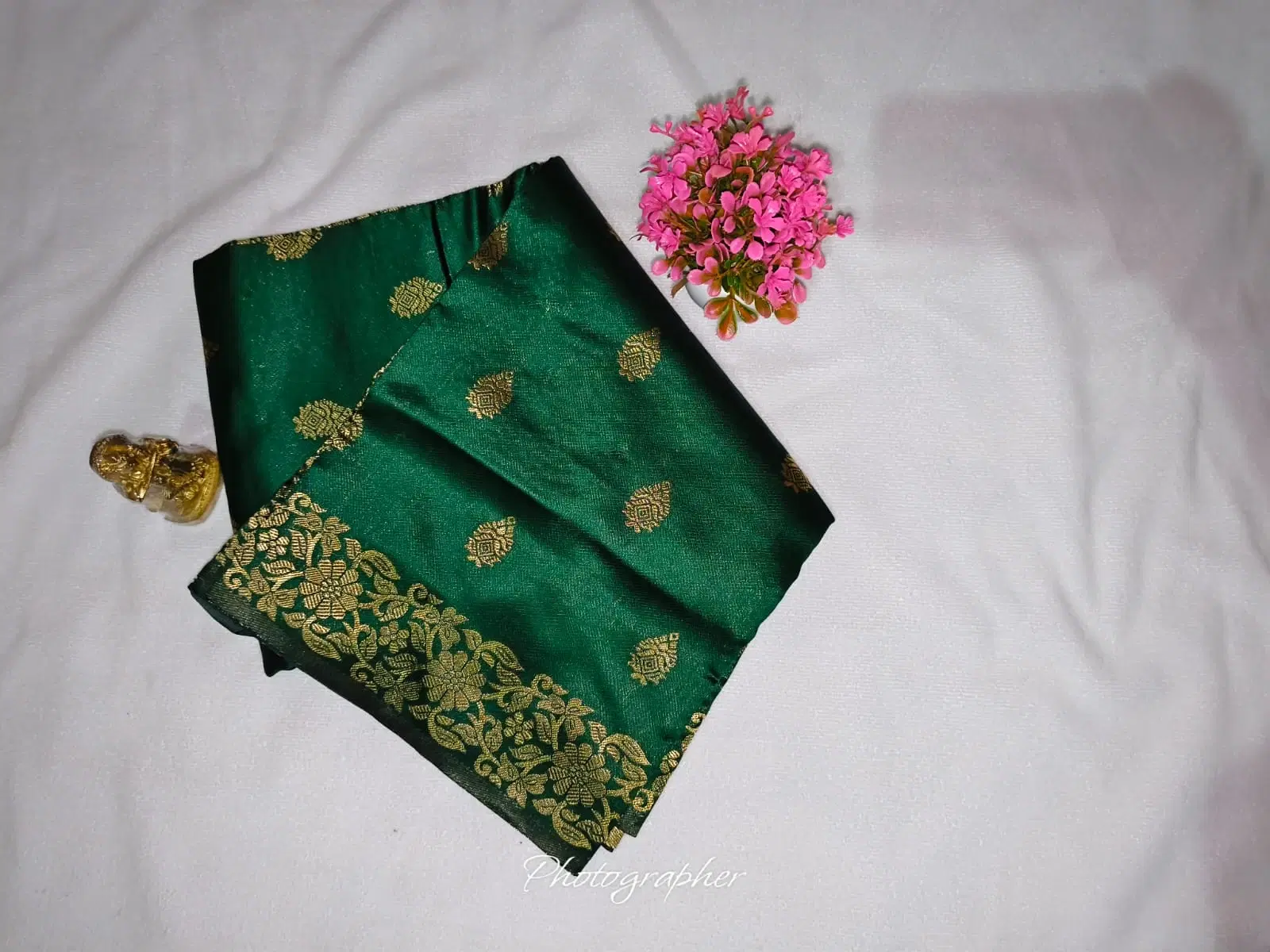Folded green silk saree with gold zari motifs and floral border on white fabric beside pink flowers