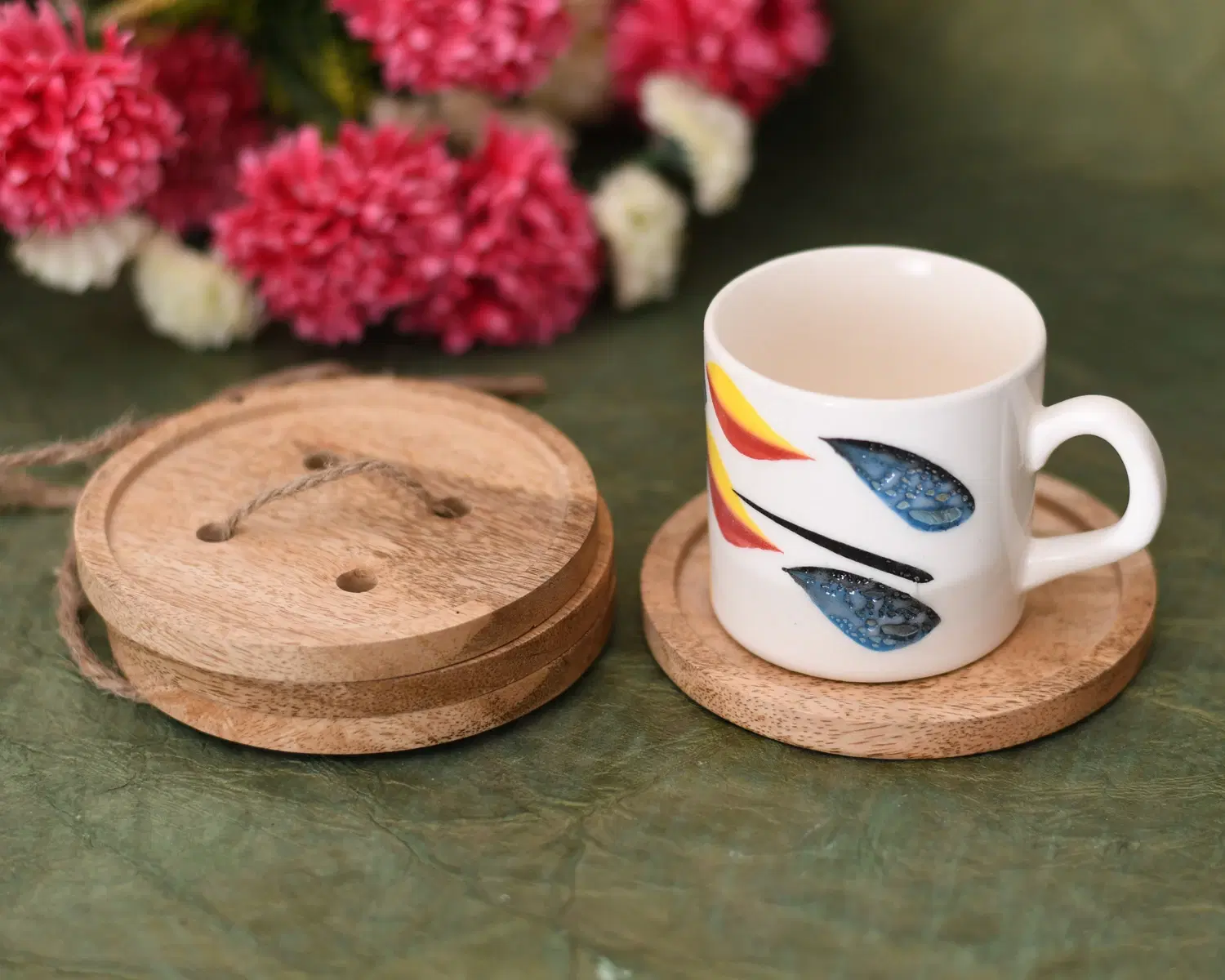 Round wooden coaster set beside a white cup with colorful leaf design on a green tabletop