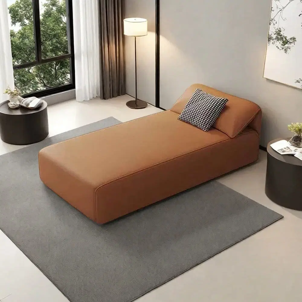 Angled front view of a brown faux leather chaise lounge on a gray rug with a checkered accent pillow