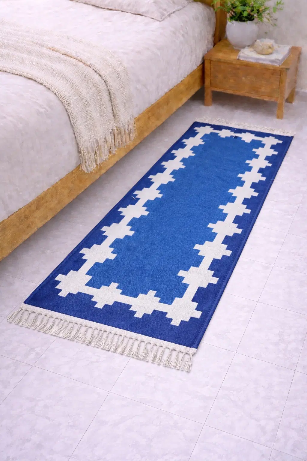 Blue runner rug with white geometric border and fringe, placed beside a wooden bed and nightstand
