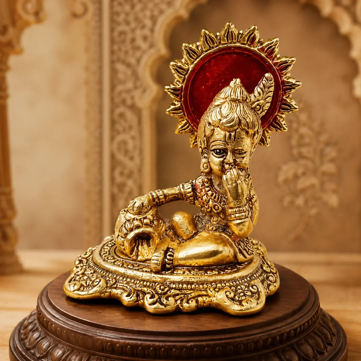 Front close view of golden Baby Krishna idol with red halo, seated on an ornate carved base
