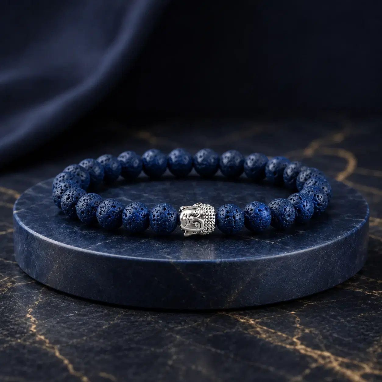 Blue porous bead bracelet with silver Buddha charm displayed on a round dark stone pedestal