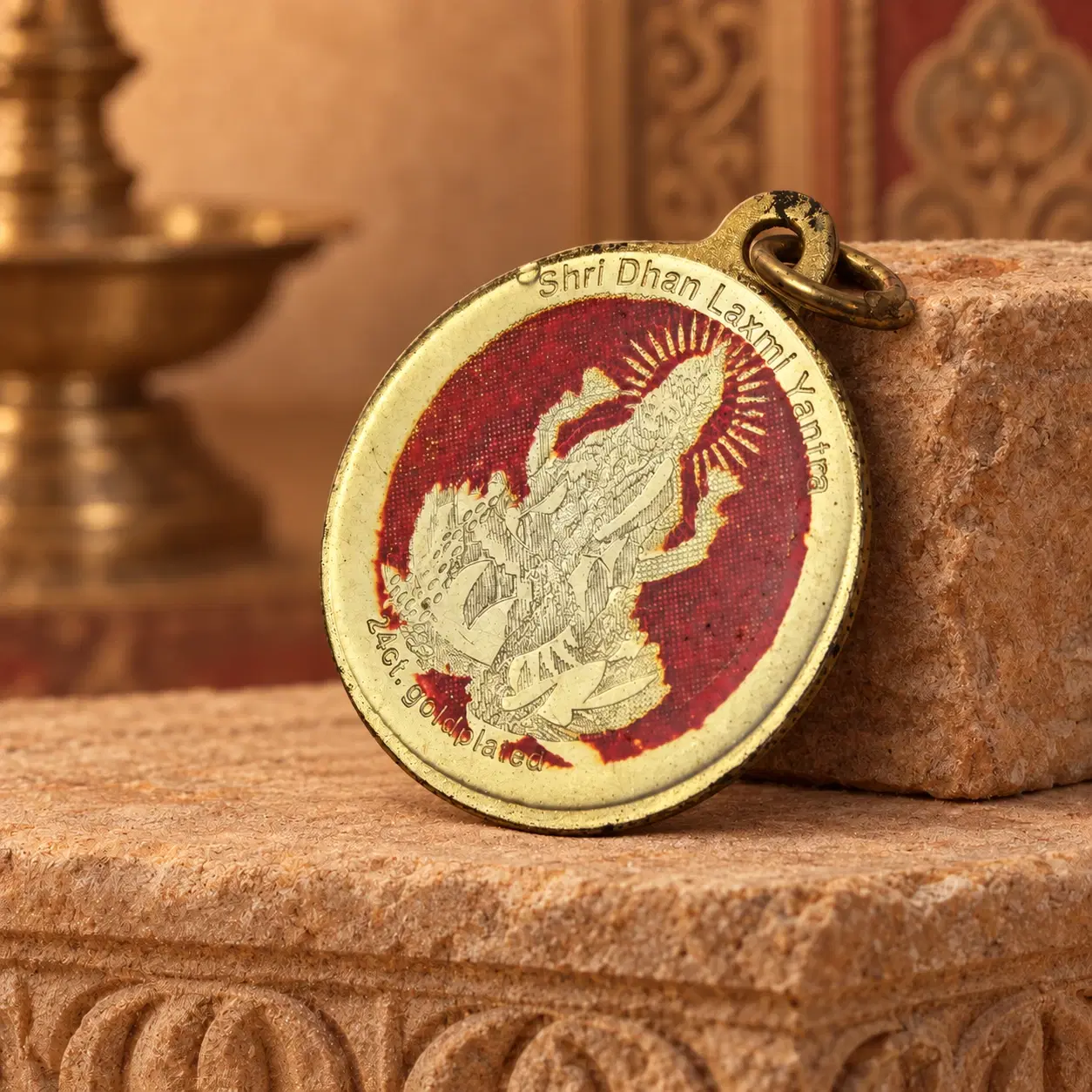 Round gold-plated Shri Dhan Laxmi Yantra pendant leaning on a stone block with red deity artwork