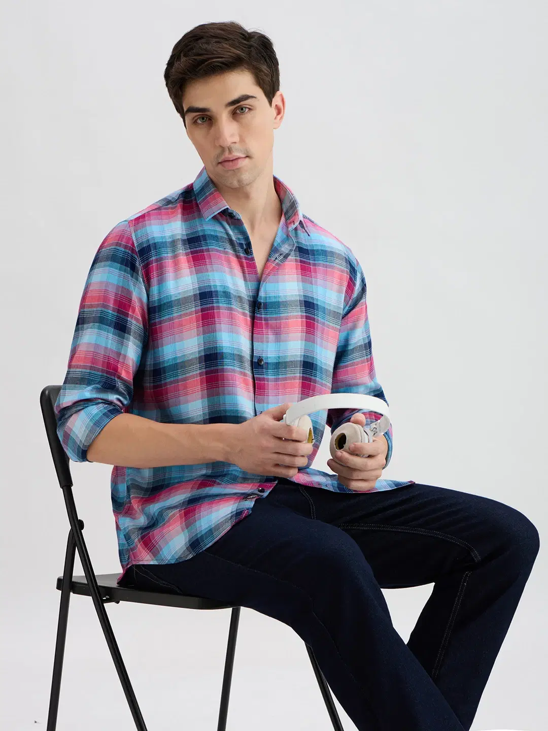 Seated angled view showing the plaid shirt drape while model holds white headphones
