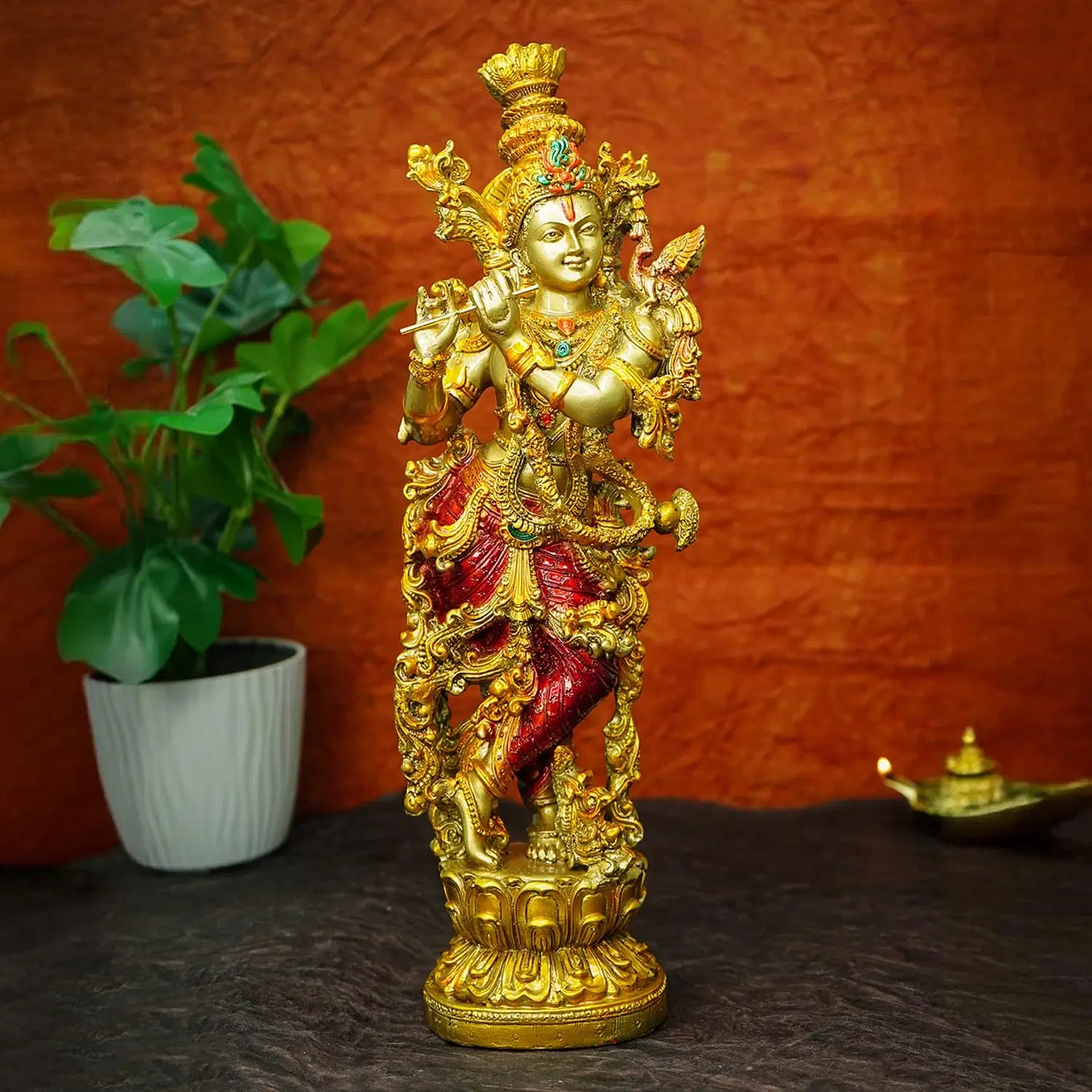 Ornate golden Lord Krishna idol with red accents standing on a pedestal beside a potted plant
