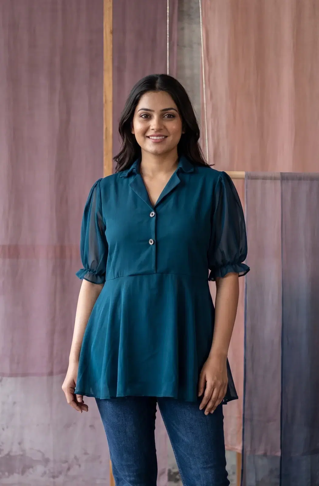 Woman wearing a teal collared tunic top with sheer puff sleeves and button detail over blue jeans