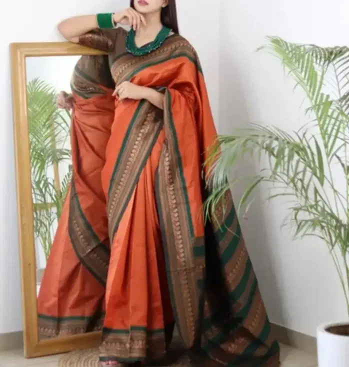 Woman wearing an orange silk saree with green and brown woven border, standing beside a mirror
